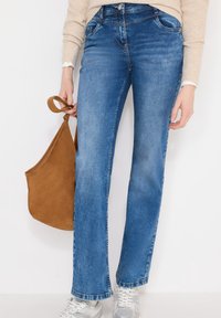 Personne portant un jean bleu, un pull beige, des baskets blanches, tenant un sac en daim marron par sa bandoulière sur un fond blanc uni.