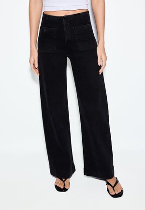 Žena nosiace čierne široké nohavice s vysokým pásom a prednými vreckami so zapínaním na gombíky, v kombinácii s čiernymi šľapkami a krátkym bielym topom.