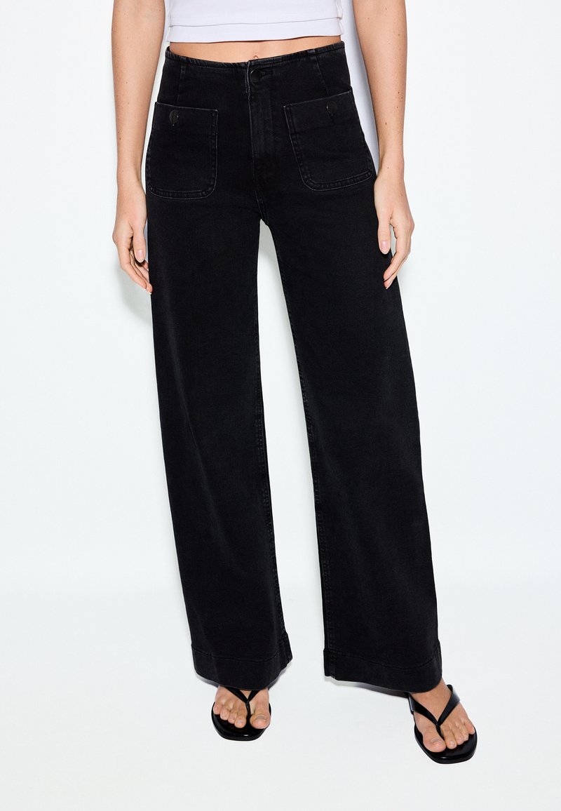 Žena nosiace čierne široké nohavice s vysokým pásom a prednými vreckami so zapínaním na gombíky, v kombinácii s čiernymi šľapkami a krátkym bielym topom.