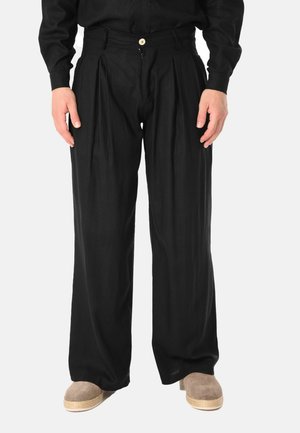 Persona che indossa pantaloni neri larghi plissettati, camicia nera a maniche lunghe e scarpe beige senza lacci su sfondo chiaro uniforme.