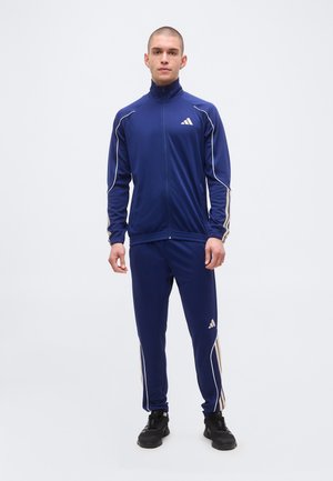 Muž stojaci oblečený v tmavomodrej Adidas súprave s bielymi pruhmi a čiernymi teniskami, otočený smerom dopredu na jednotnom svetlom pozadí.