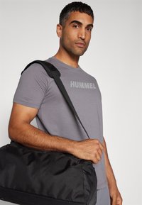 Camiseta de manga corta gris con la impresión "HUMMEL", combinada con una gran bolsa de deportes negra que cuenta con una correa resistente y una textura suave.