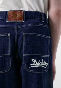 Tamsiai mėlynos džinsinės kelnes su odiniu žymekliu ant juosmens ir baltu siuvinėtuo "Dickies" logotipu dešinėje užpakalinėje kišenėje.