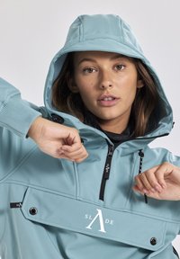 Giacca a vento azzurro chiaro con cappuccio, tasca frontale, cordini regolabili e logo. Realizzata in tessuto liscio e opaco con polsini elasticizzati.