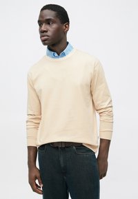 Beež puuvillane dressipluus ümar kaelusega, pikkade varrukatega ning selle all hele sinine kraega särk; tumesinised teksapüksid ja pruun vöö.