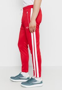 Rode sportieve broek met witte zijstrepen, voorzien van een elastische tailleband en een gladde, synthetische textuur. Gecombineerd met turquoise sneakers.