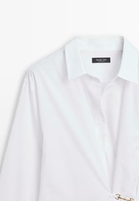 Chemise blanche à manches longues avec col et patte de boutonnage cachée, ornée d’un petit fermoir doré près de la taille, étiquetée Massimo Dutti.