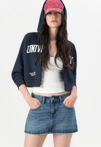 Sudadera navy corta con "UNIVERSITY" impreso en blanco, combinada con una camiseta sin mangas blanca y una falda mini de denim con dobladillo deshilachado y bolsillos.