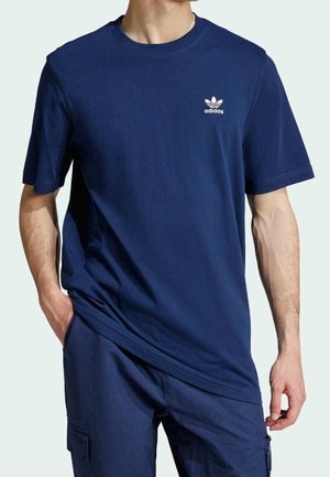 Homme portant un t-shirt Adidas bleu marine avec logo sur la poitrine et un pantalon cargo bleu marine, une main dans la poche, sur fond clair.