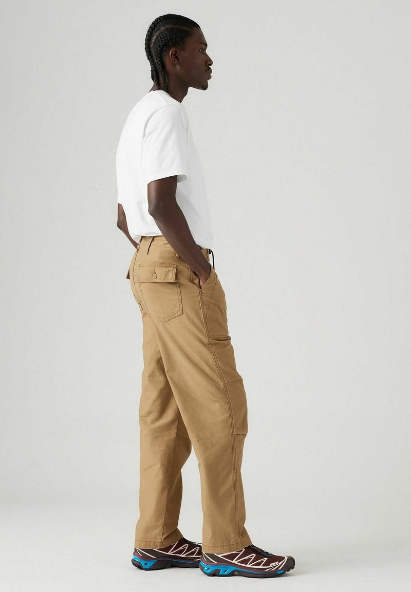 Pantalones cargo beige con bolsillos delanteros y traseros, llevados con una camiseta blanca y zapatillas multicolores. Textura suave, corte relajado.