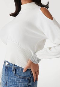 Pulover alb cu guler rib, umeri decupați și manșete accentuate cu argintiu. Asortat cu blugi albaștri cu dungi verticale argintii.