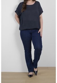 Donkergrijze, short-sleeve top van lichtgewicht stof, gecombineerd met donkerblauwe slim-fit jeans en puntige zwarte hakken. Eenvoudige achtergrond.