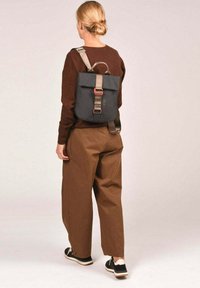 Mochila de tela negra con cierre superior plegable, correas beige y detalles en rojo. Se lleva puesta en la espalda, combinada con pantalones anchos marrones.