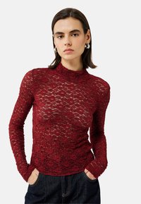 Top lungo a maniche lunghe in pizzo rosso con collo alto, design aderente e intricato motivo floreale. Indossato con jeans di denim blu scuro.