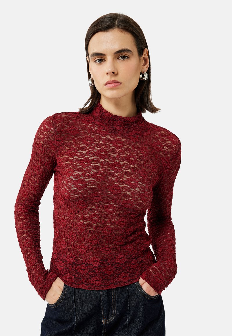 Top largo de encaje rojo con mangas largas y un cuello alto, diseño ajustado y patrón floral intrincado. Combinado con jeans de mezclilla azul oscuro.