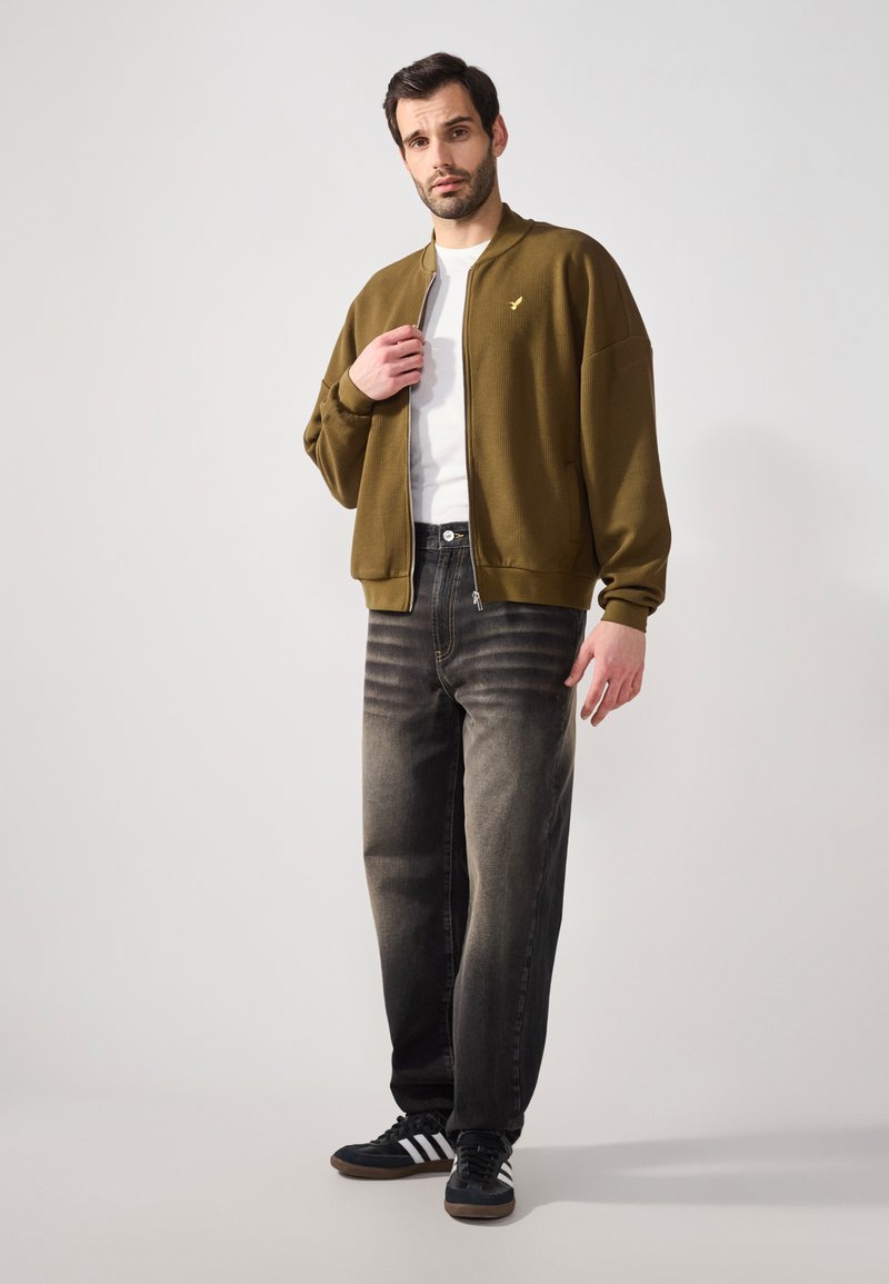 Olivgrüne Bomberjacke aus strukturiertem Stoff, weißes T-Shirt, ausgebleichte schwarze Jeans und schwarze Sneakers mit weißen Streifen. Goldenes Logo auf der Jacke.