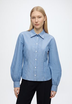 Femme portant un chemisier à rayures verticales bleues et blanches avec manches longues bouffantes et pantalon noir, debout devant un fond blanc.