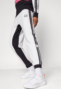 Šedé a černé tepláky mají zúžený střih, kontrastní boční panely a logo na stehně. Jsou kombinovány s bílými sportovními botami.