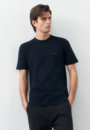 Νεαρός άνδρας με σκούρα μαλλιά, φορώντας μαύρο κοντομάνικο πουκάμισο με το λογότυπο "BOSS", στέκεται με τα χέρια στις τσέπες μπροστά σε λευκό φόντο.