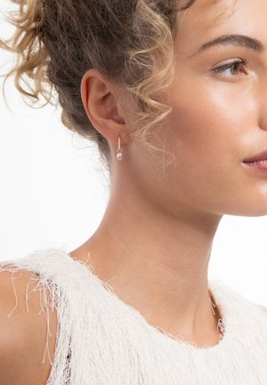 Boucle d'oreille en or en forme d'anneau avec un petit détail de perle. Le modèle porte un vêtement blanc texturé avec des accents à franges.