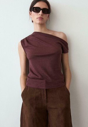 Frau mit dunkler Sonnenbrille, einem ein-schulterigen bordeauxfarbenen Top und braunen hoch taillierten Hosen, die vor einer einfachen hellen Wand steht.