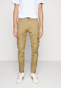 Ljust bruna cargo-byxor med fickor på båda benen, tillverkade av bomullstyg, med bältesöglor och knappstängning, i kombination med vita sneakers.