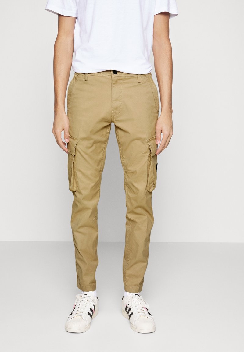 Ljust bruna cargo-byxor med fickor på båda benen, tillverkade av bomullstyg, med bältesöglor och knappstängning, i kombination med vita sneakers.