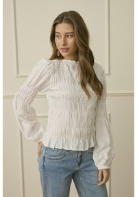 Junge Frau mit langem, welligem Haar, die eine weiße smockartige Bluse und blaue Jeans trägt, steht mit einer Hand in der Hüfte und schaut zur Seite.