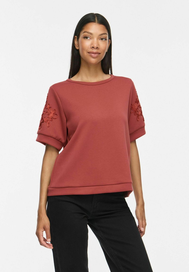 Femme portant un haut court à manches courtes couleur rouille avec broderies florales sur les manches et un pantalon noir, debout devant un fond blanc uni.