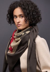 Sciarpa marrone scuro e beige con accenti rossi, caratterizzata da un motivo geometrico. Indossata sovrapposta a una giacca beige chiaro. Texture morbida.