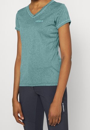 Osoba ubrana w krótki turkusowy T-shirt z dekoltem w serek i ciemne sportowe spodnie z logo na udzie, stojąca na jednolitym tle.