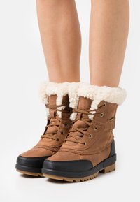 Botas impermeables marrones con suela de goma negra, adornadas con un borde de piel de oveja en la parte superior, diseño de cordones y paneles de cuero texturizado.