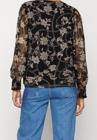 Blusa nera trasparente con motivo floreale in beige. Maniche lunghe a sbuffo con polsini arricciati. Tessuto leggero. Abbinata a jeans in denim blu chiaro.