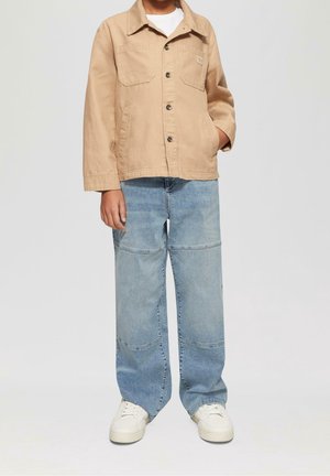 Chaqueta de algodón marrón con cierre de botones y dos bolsillos en el pecho, combinada con jeans de parcheado azul claro y zapatillas blancas.