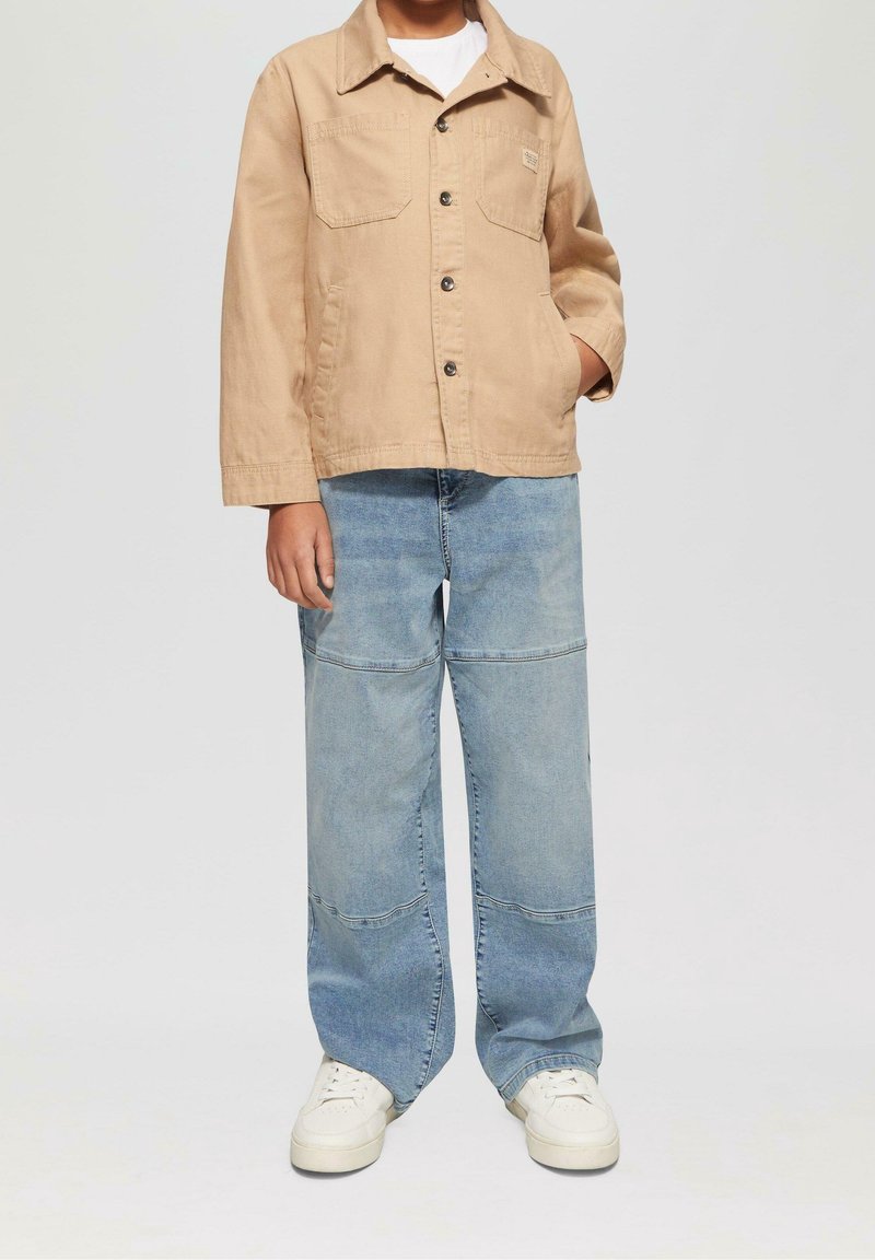 Chaqueta de algodón marrón con cierre de botones y dos bolsillos en el pecho, combinada con jeans de parcheado azul claro y zapatillas blancas.