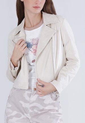Donna con giacca in camoscio beige chiaro sopra una maglietta bianca con stampa floreale e pantaloni chiari con motivo, che sistema la cerniera della giacca.