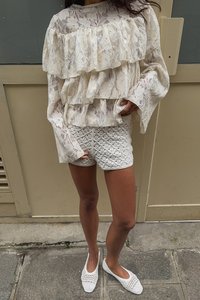 Haut en dentelle crème avec des volants superposés, associé à un short en crochet blanc. Porté avec des chaussures slip-on tissées blanches, dans un cadre urbain.