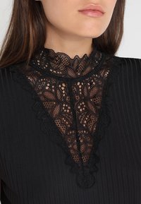 Haut noir avec un col en dentelle haute présentant des motifs floraux et des bords à volants, fabriqué en tissu texturé avec des nervures verticales.