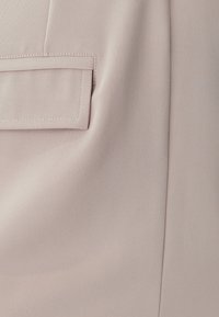 Světle růžová látka s jemnou strukturou obsahuje malou šikmou kapsu a viditelné stehy. Materiál vypadá hladce a elegantně.