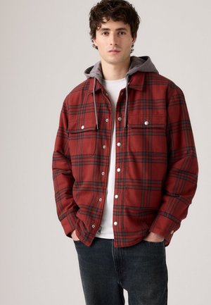 Red plaid flannel shirt with grey hood, snap buttons, two chest pockets, and textured fabric. Worn over a white t-shirt and dark jeans.