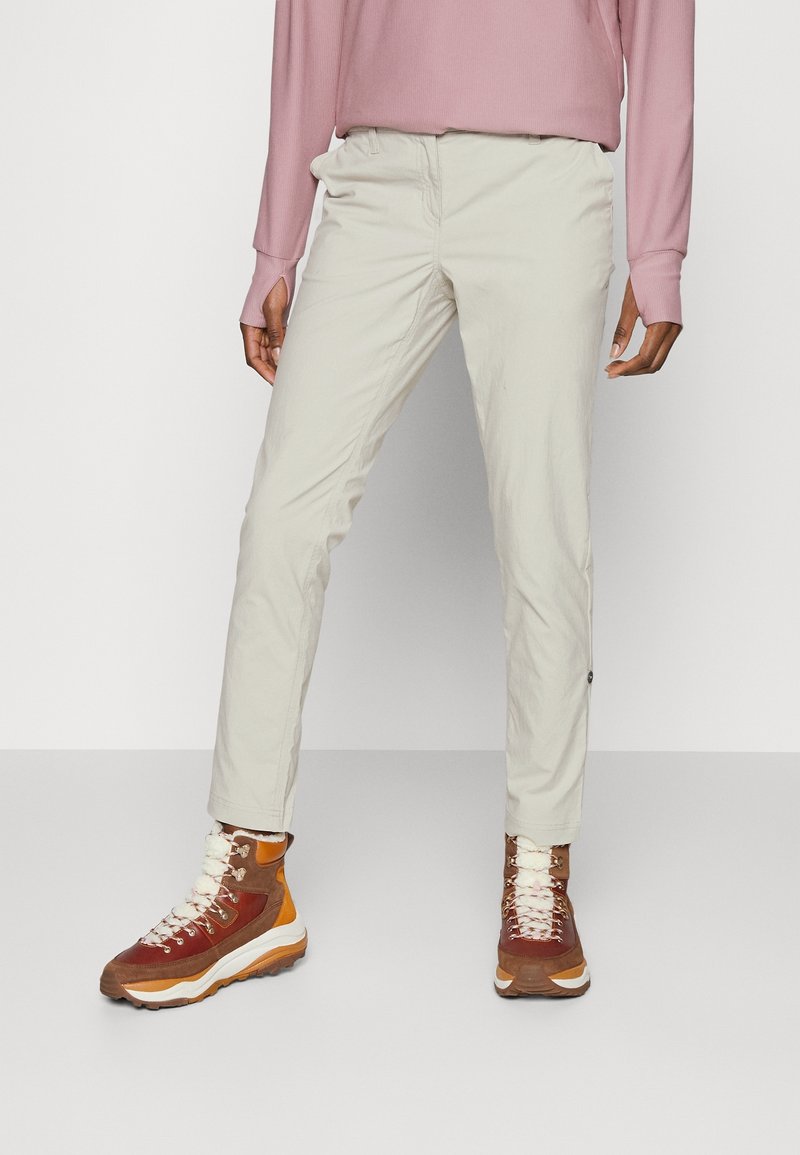 Personne portant un pantalon beige, un haut rose à manches longues et des bottes de randonnée marron, debout sur un sol gris clair uni.