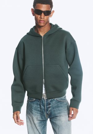 Jeune homme portant des lunettes de soleil noires, un sweat à capuche vert foncé à fermeture éclair et un jean bleu clair, debout les mains détendues le long du corps.