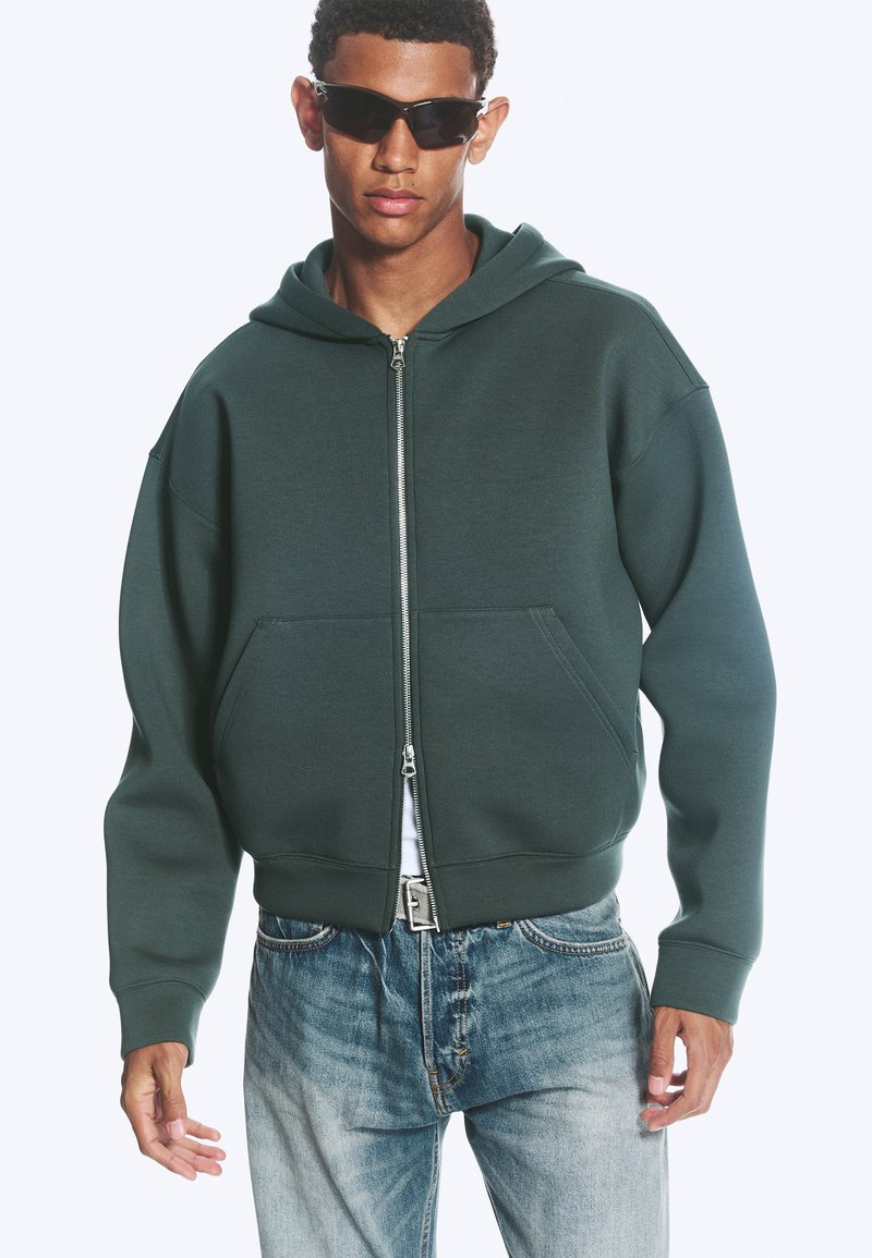 Jeune homme portant des lunettes de soleil noires, un sweat à capuche vert foncé à fermeture éclair et un jean bleu clair, debout les mains détendues le long du corps.