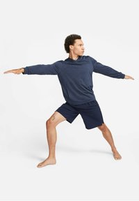 Navy blue long-sleeve hoodie with a textured finish, paired with dark shorts. The figure poses in a wide stance, barefoot, with visible tattoo.