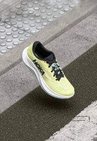 Hoka hardloopschoen met een lichtgroene mesh bovenkant, zwarte accenten, dikke witte tussenzool en blauwe buitenzool, ontworpen voor comfort en ondersteuning.