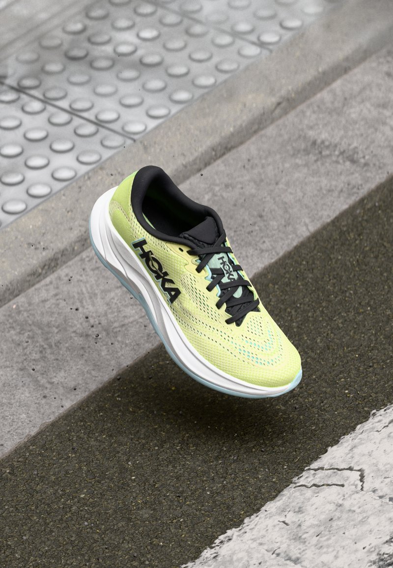 Hoka hardloopschoen met een lichtgroene mesh bovenkant, zwarte accenten, dikke witte tussenzool en blauwe buitenzool, ontworpen voor comfort en ondersteuning.