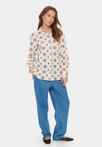 Bedruckte Bluse in Creme mit geometrischen Mustern in Burgunderrot und Marineblau. Kombiniert mit locker sitzenden blauen Jeans. Schwarze Ballerinas.