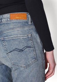 Jeans de mezclilla de color azul claro con una etiqueta de cuero marrón con "REPLAY" grabado. El bolsillo trasero incluye un diseño de costura azul ondulada.