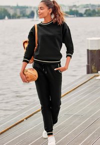 Schwarzer Sportpullover mit weißen Akzenten, kombiniert mit schwarzen Jogginghosen und weißen Sneakers. Hält einen Basketball und einen braunen Rucksack.