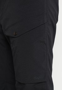 Pantalons cargo noirs avec une finition texturée, dotés d'une poche à rabat diagonale fermée par un bouton et d'un logo brodé près de l'ourlet.